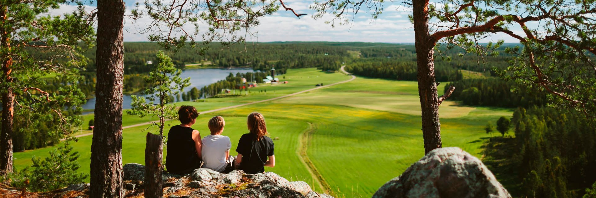 Three people sit on a hill (Falkberget) and look out over a cultivated landscape with a lake.