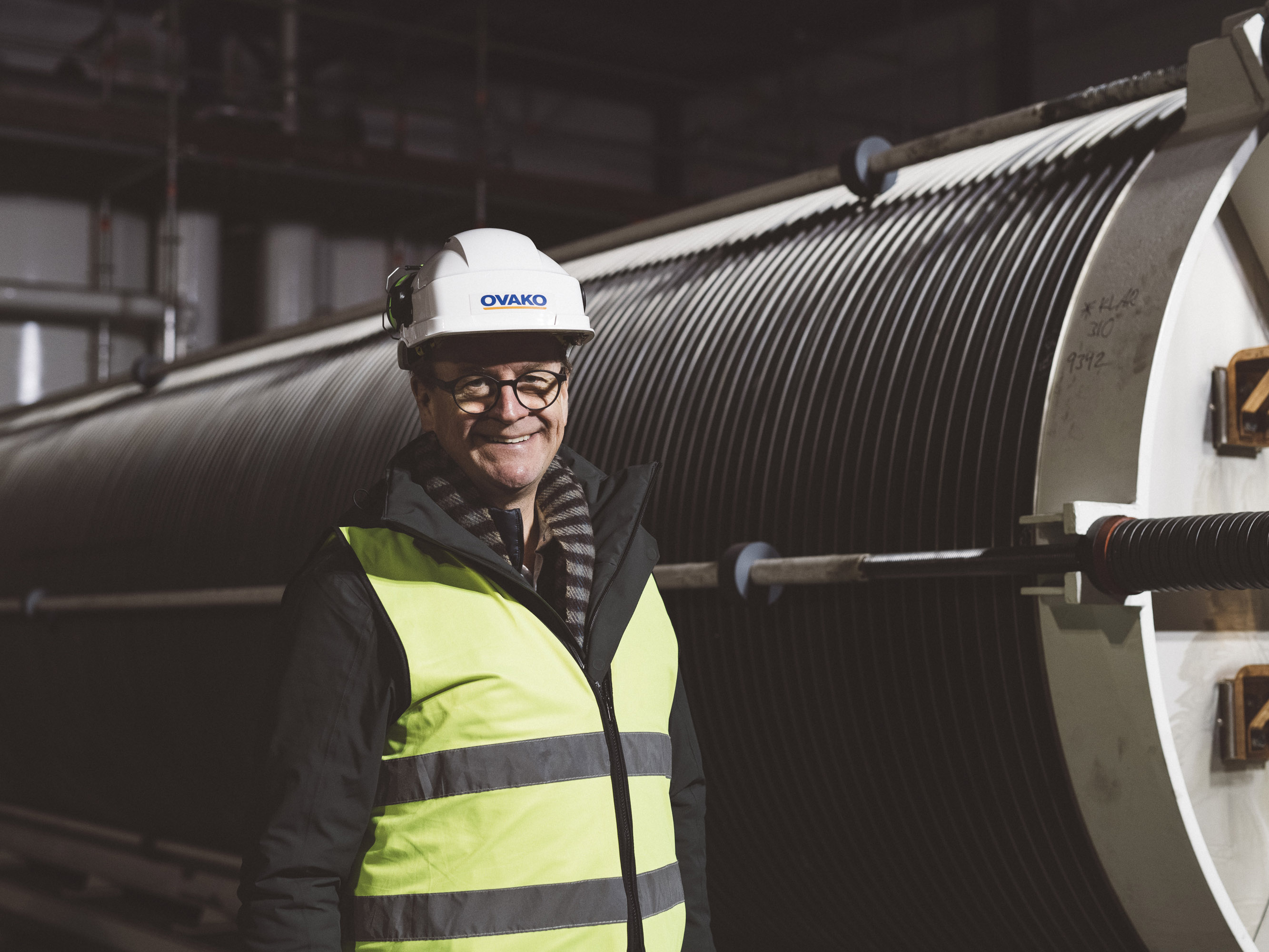 Man med glasögon, hjälm och varselväst i industrimiljö.