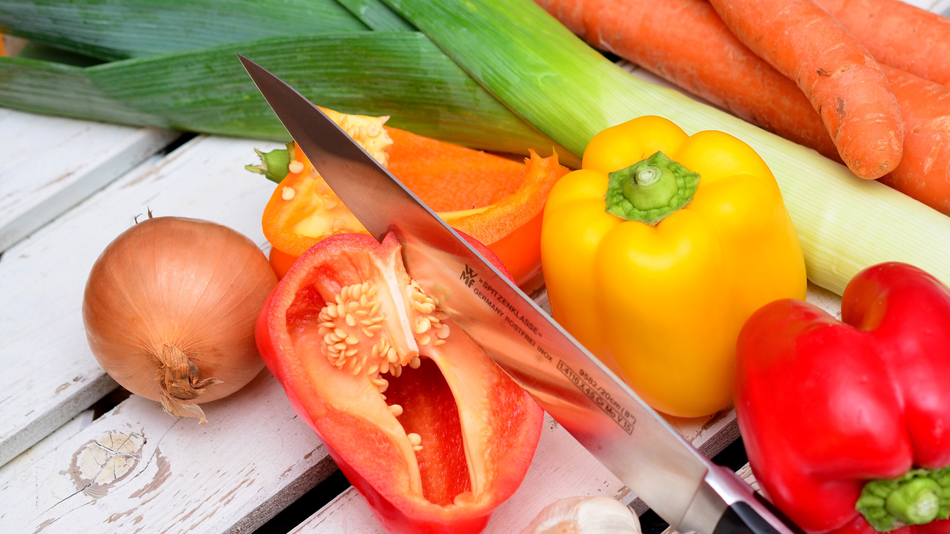 En kniv skär genom en paprika och andra grönsaker ligger runtomkring.