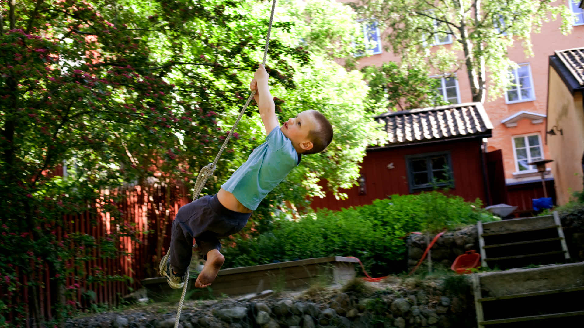 Pojke som hänger i ett rep på i grönskande förskolegård.